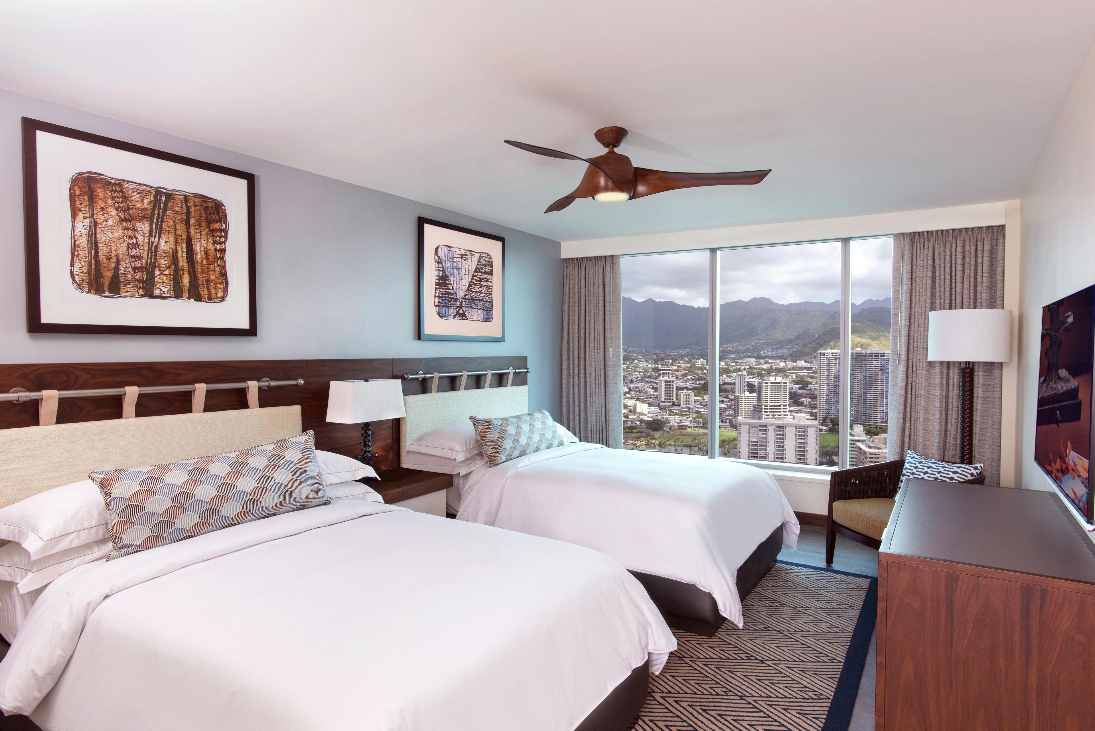 A hotel room with two beds, a ceiling fan, large windows with a city and mountain view, and artwork on the walls.