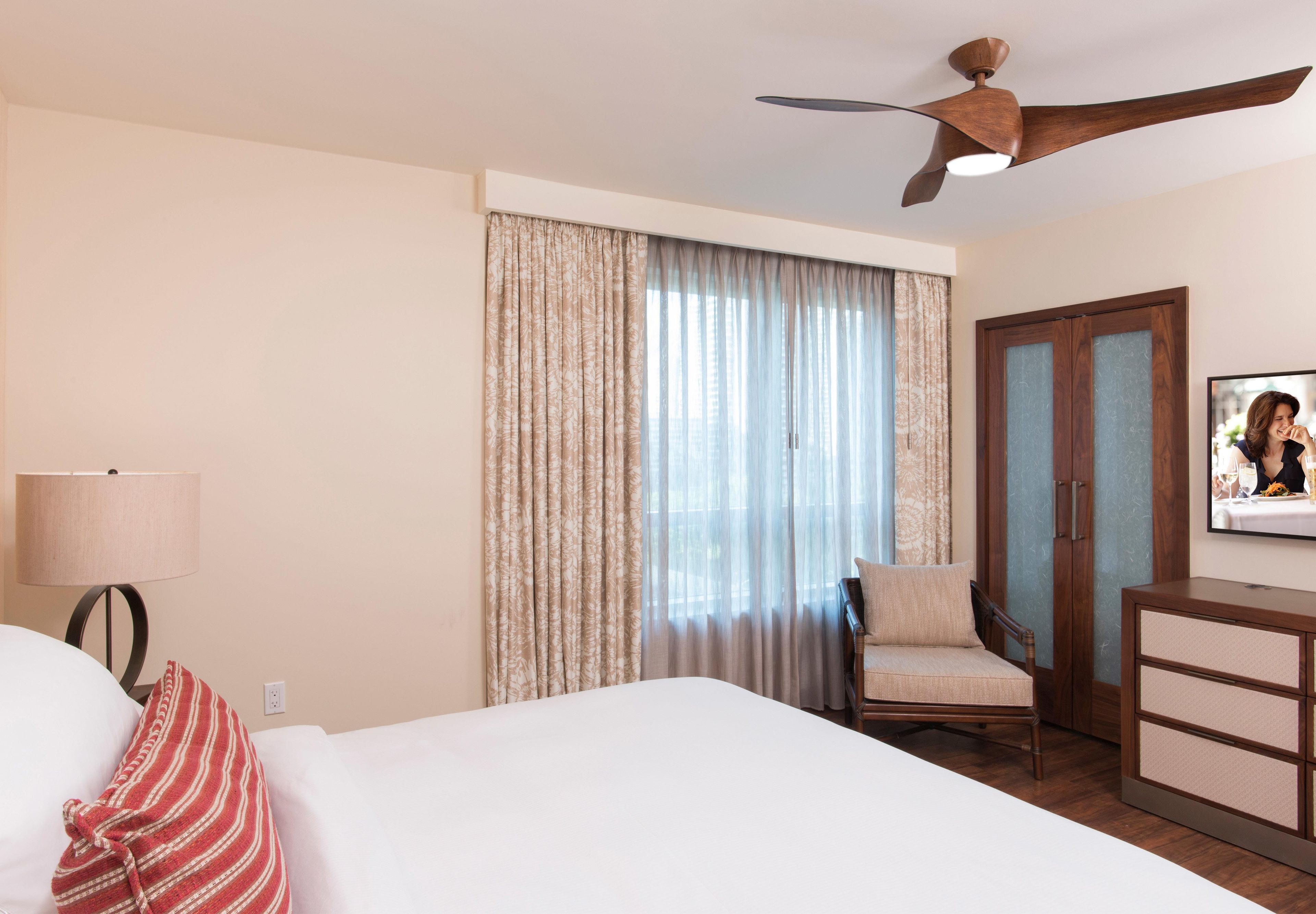 Ein Hotelzimmer mit Bett, Sessel, Schreibtisch, Deckenventilator und einem Fenster mit Vorhängen.