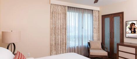 Ein Hotelzimmer mit Bett, Sessel, Schreibtisch, Deckenventilator und einem Fenster mit Vorhängen.