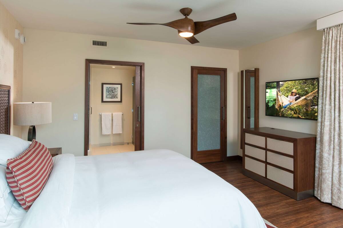 Ein Hotelzimmer mit einem Bett, einem Fernseher, einem Deckenventilator, einem Schminktisch und einer Tür zu einem Badezimmer.