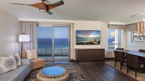 A hotel room with a large sliding glass door to the ocean, a dining area with a table and chairs, a sofa with patterned cushions, a round wicker ottoman, and a ceiling fan. - Hilton Grand Vac Club The Grand Islander Waikiki Honolulu (Honolulu)