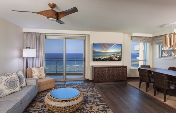 A hotel room with a large sliding glass door to the ocean, a dining area with a table and chairs, a sofa with patterned cushions, a round wicker ottoman, and a ceiling fan. - Hilton Grand Vac Club The Grand Islander Waikiki Honolulu (Honolulu)