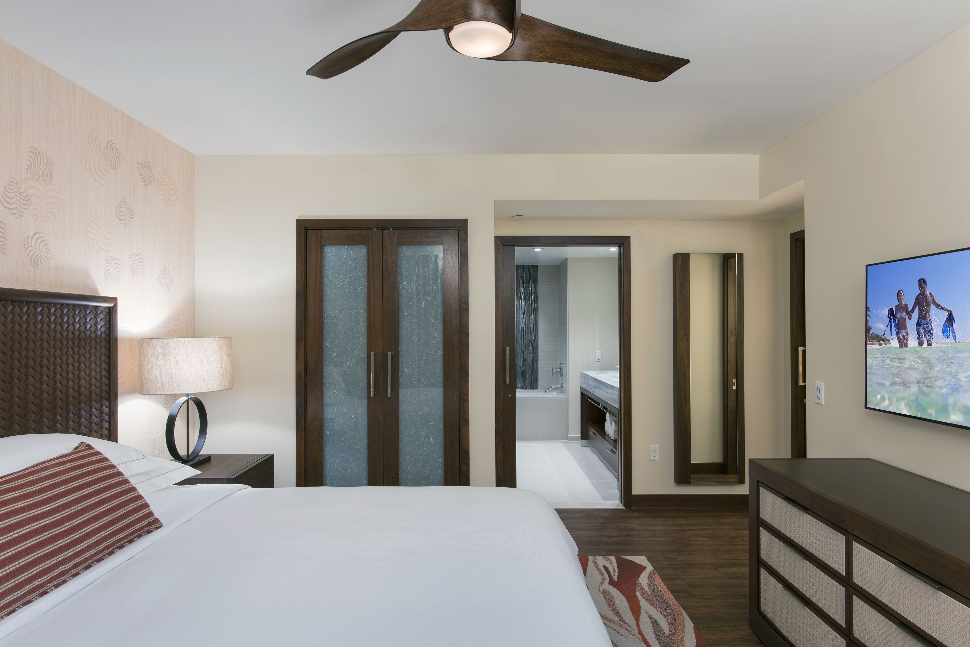 A hotel room with a bed, a ceiling fan, a TV, a bathroom with a shower, and a mirror.