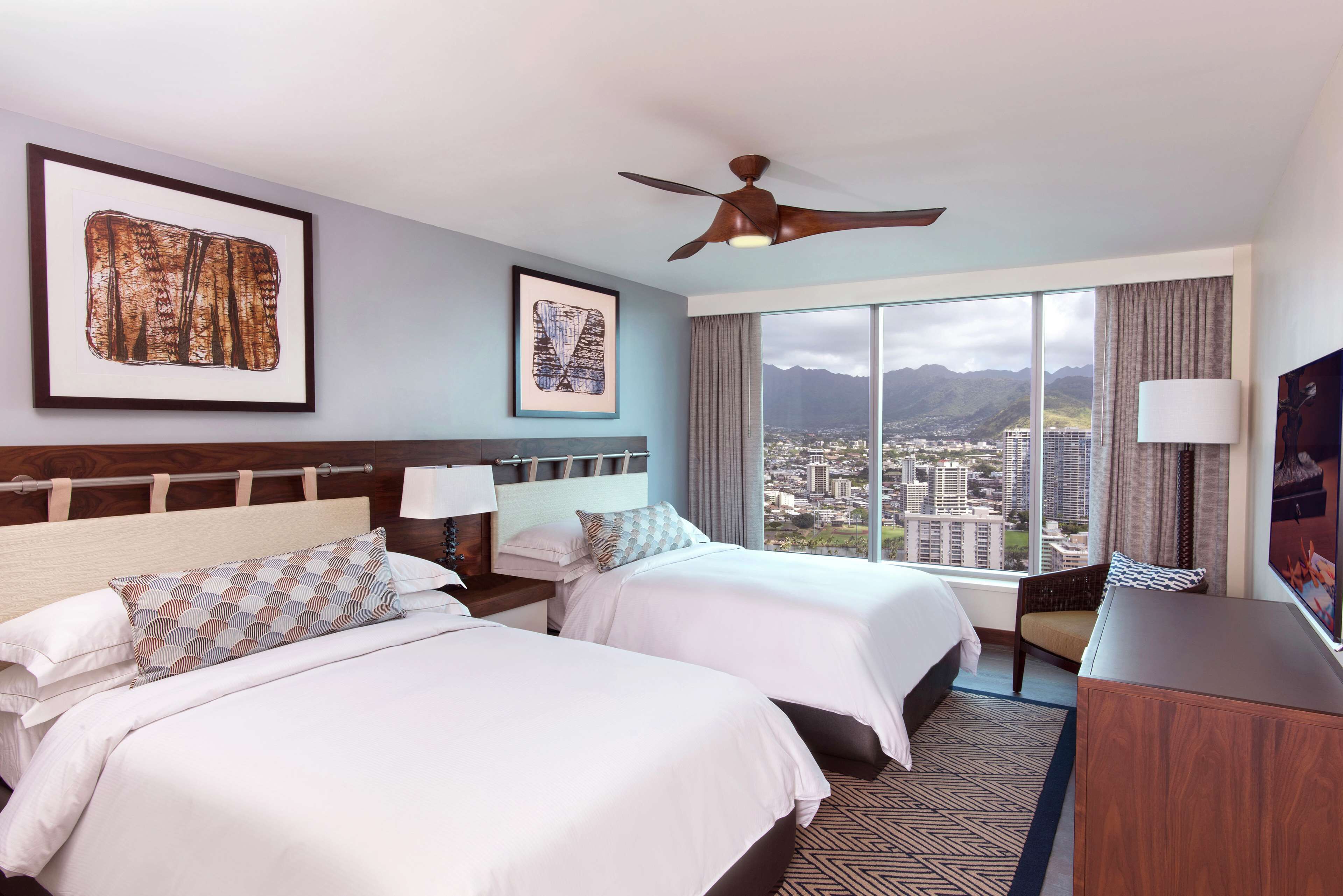 Una habitación de hotel con dos camas, ventilador de techo, ventanas amplias con vista a la ciudad y a las montañas, y obras de arte en las paredes.
