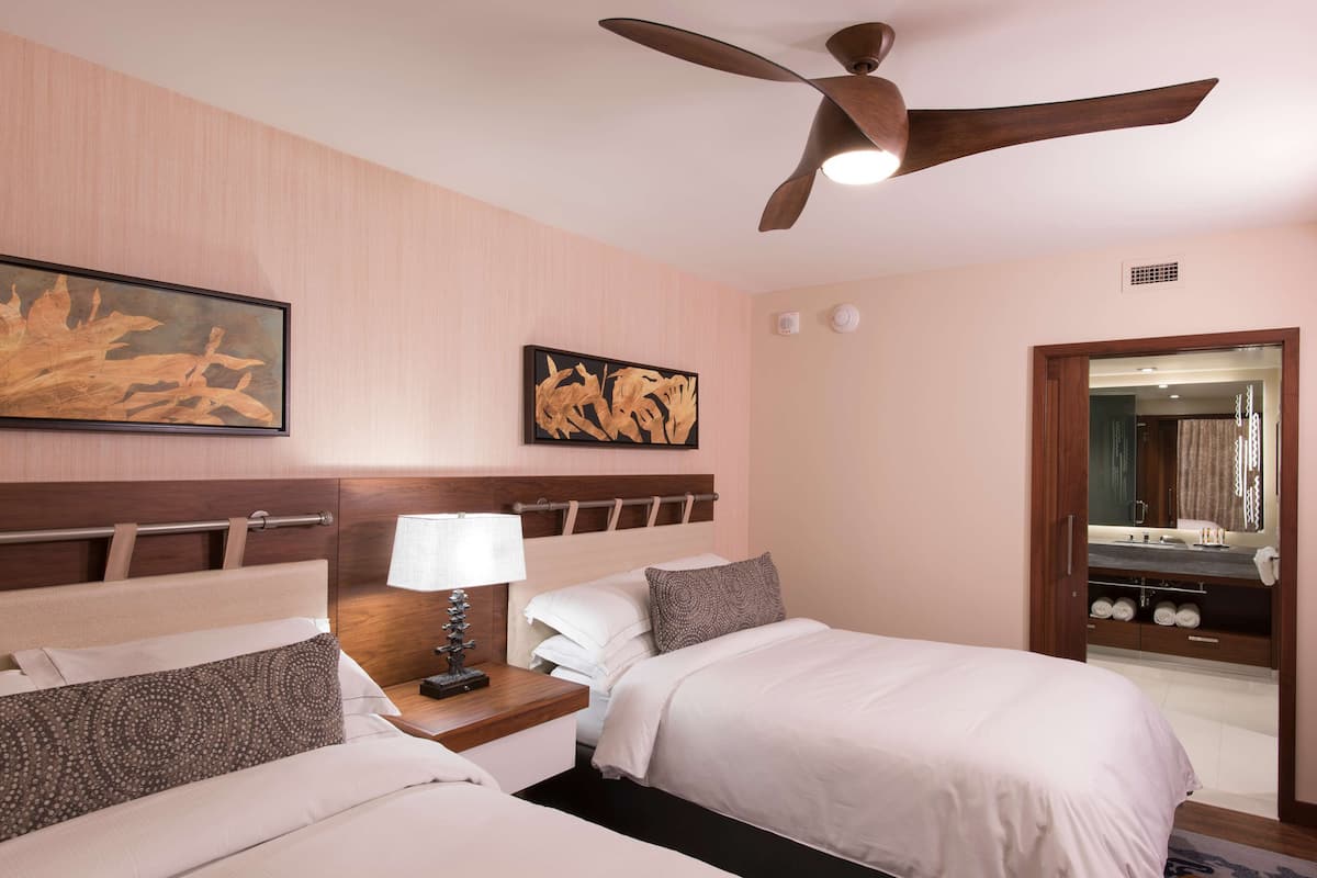 Ein Hotelzimmer mit zwei Betten, einem Deckenventilator, Bildern an den Wänden, einem Nachttisch mit Lampe und einem durchgehenden Türspalt sichtbaren Badezimmer.