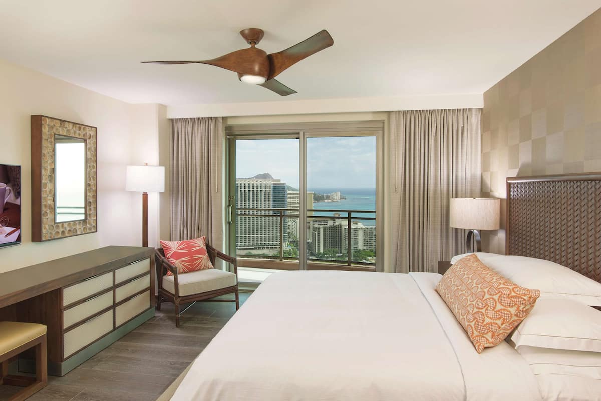 Ein Hotelzimmer mit Bett, Schreibtisch, Stuhl, Deckenventilator und einem großen Fenster mit Blick auf die Stadt.