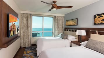 A hotel room with two beds, a ceiling fan, a large window with a view of the ocean, and artwork on the walls.