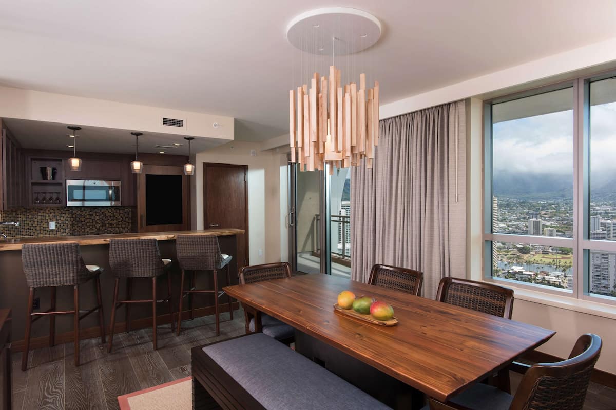 Ein moderner Essbereich mit einem Holztisch, Barhocker und einer Obstschale. Ein großes Fenster bietet Blick auf die Stadt.