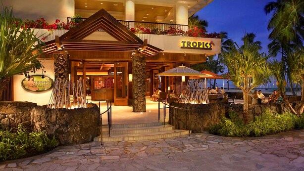 A Tropics restaurant with a stone facade, outdoor seating, and a view of the ocean. - Hilton Grand Vac Club The Grand Islander Waikiki Honolulu (Honolulu)