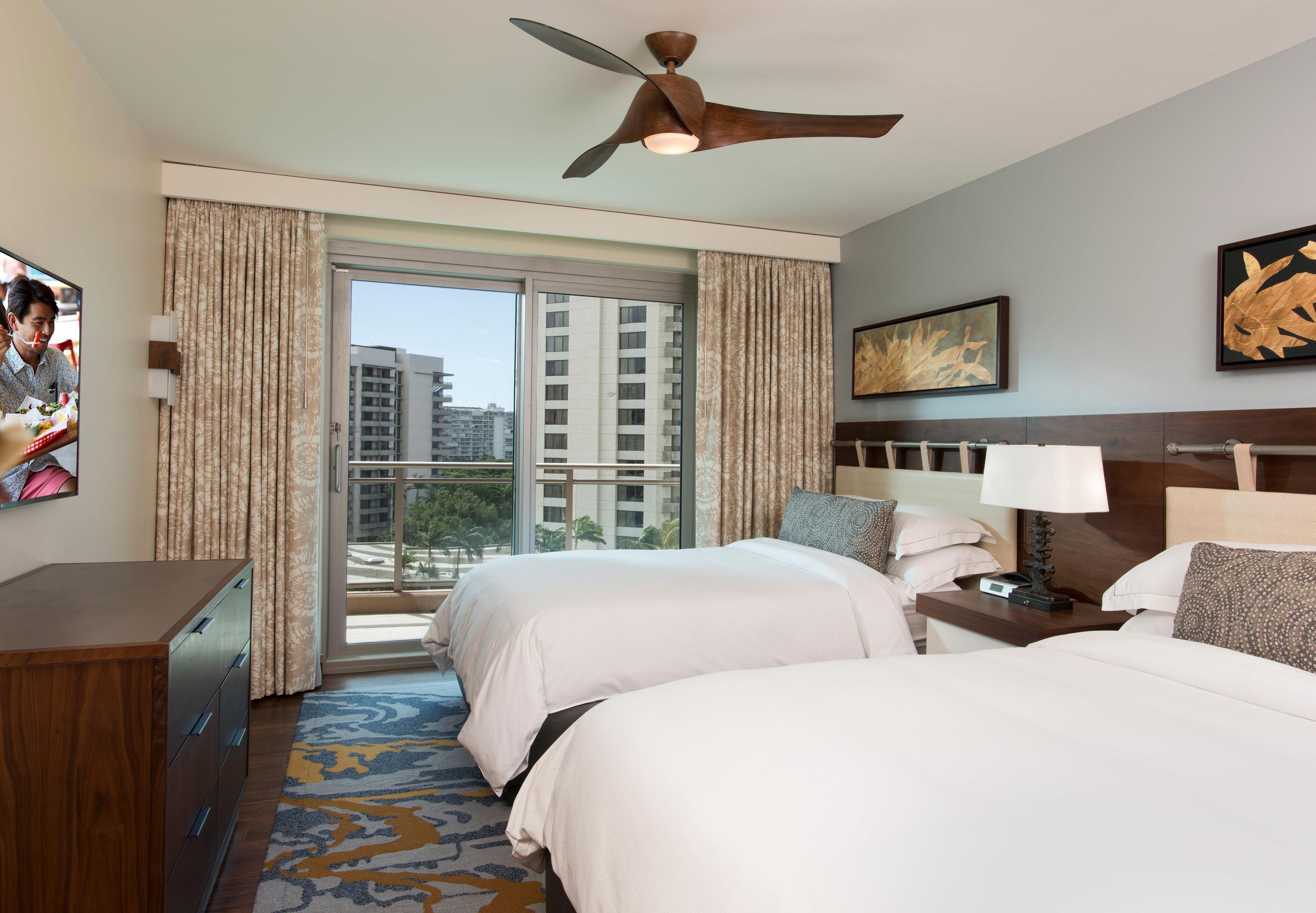 A hotel room with two beds, a ceiling fan, a window with curtains, a nightstand with a lamp, and a painting on the wall.