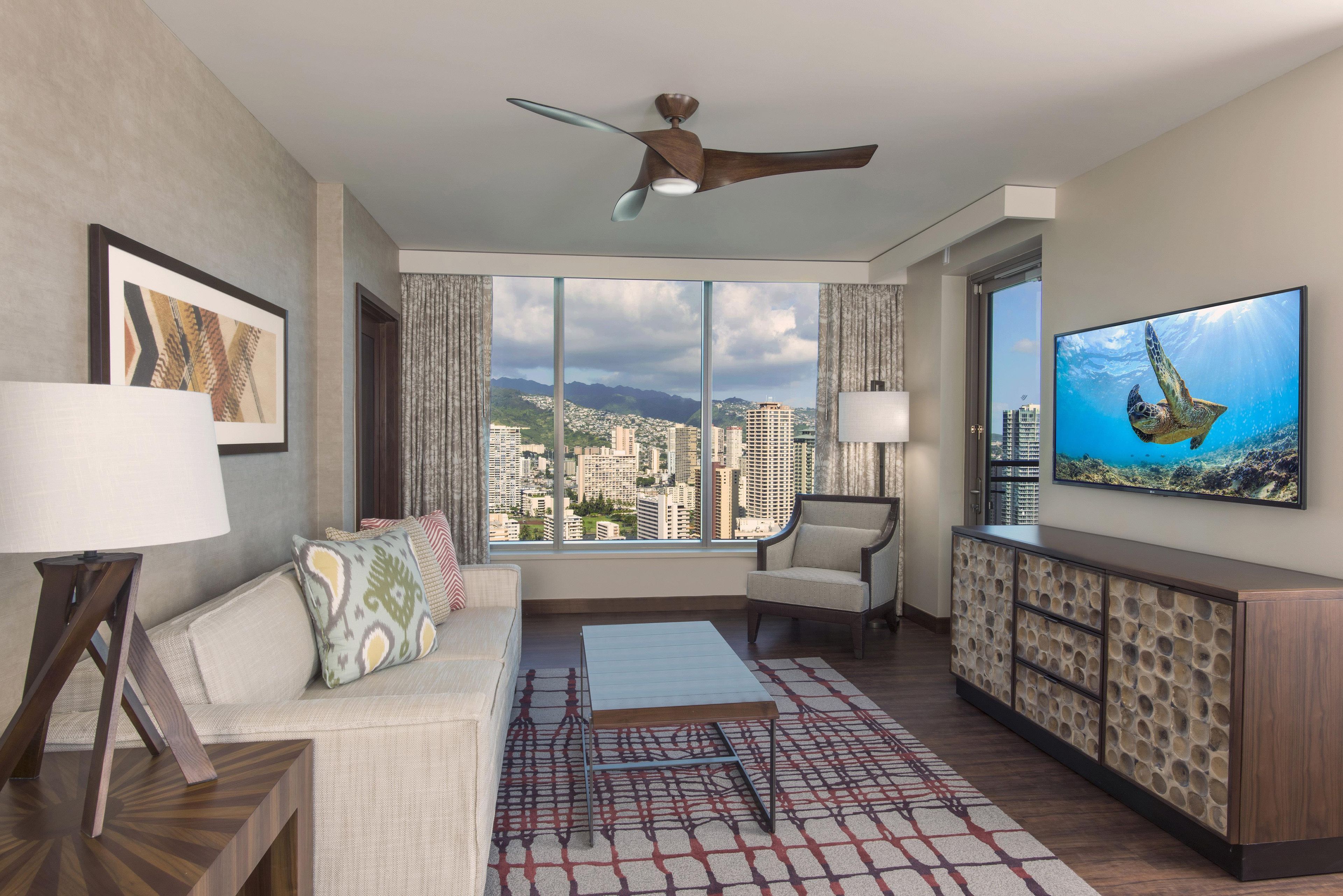 Ein modernes Hotelzimmer mit Blick auf die Stadt, inklusive Deckenventilator, Sofa, Couchtisch, Fernseher und einem gemusterten Teppich.