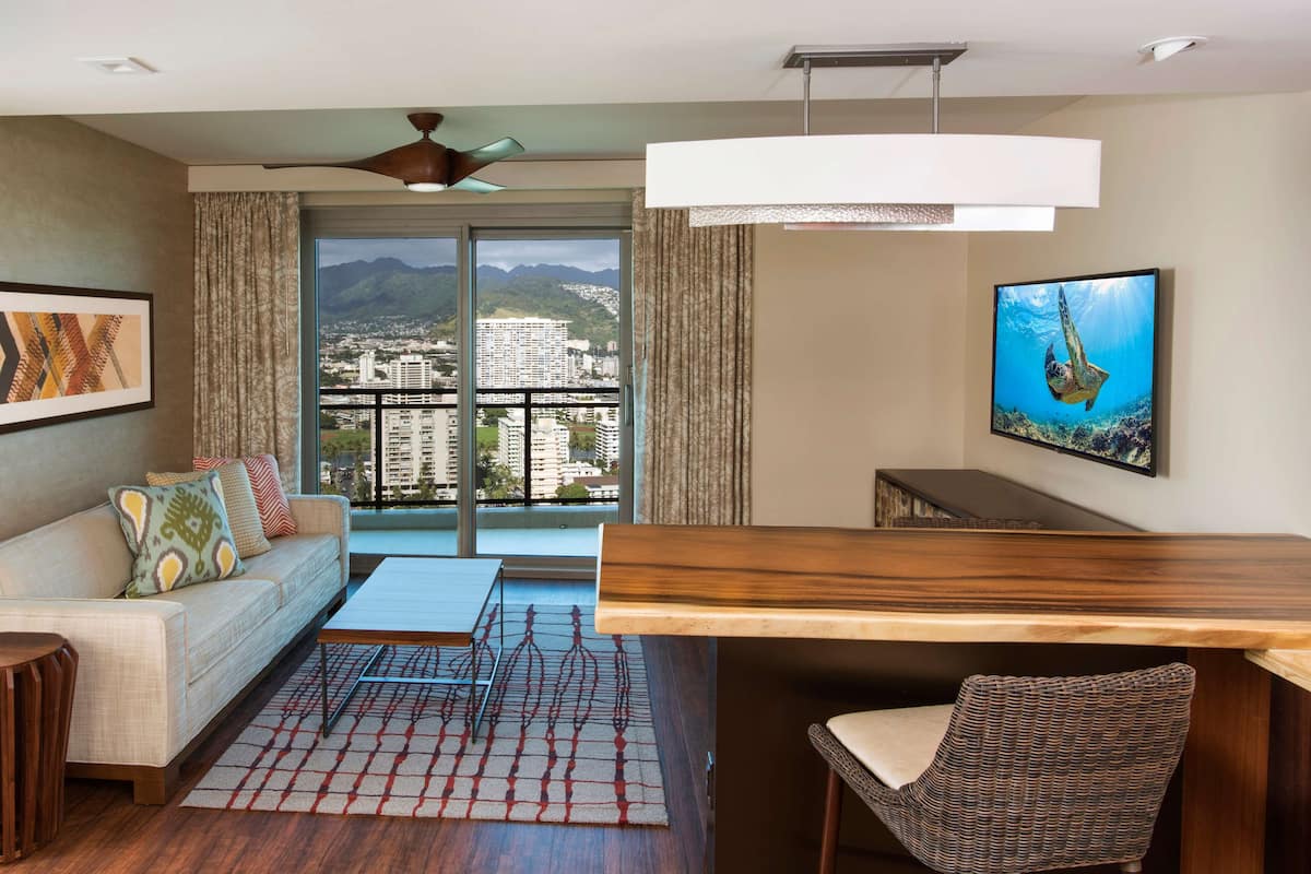 Ein modernes Hotelzimmer mit Balkon, einer Couch mit gemusterten Kissen, einem Holztisch und einem Fernseher, der eine Unterwasserlandschaft zeigt.