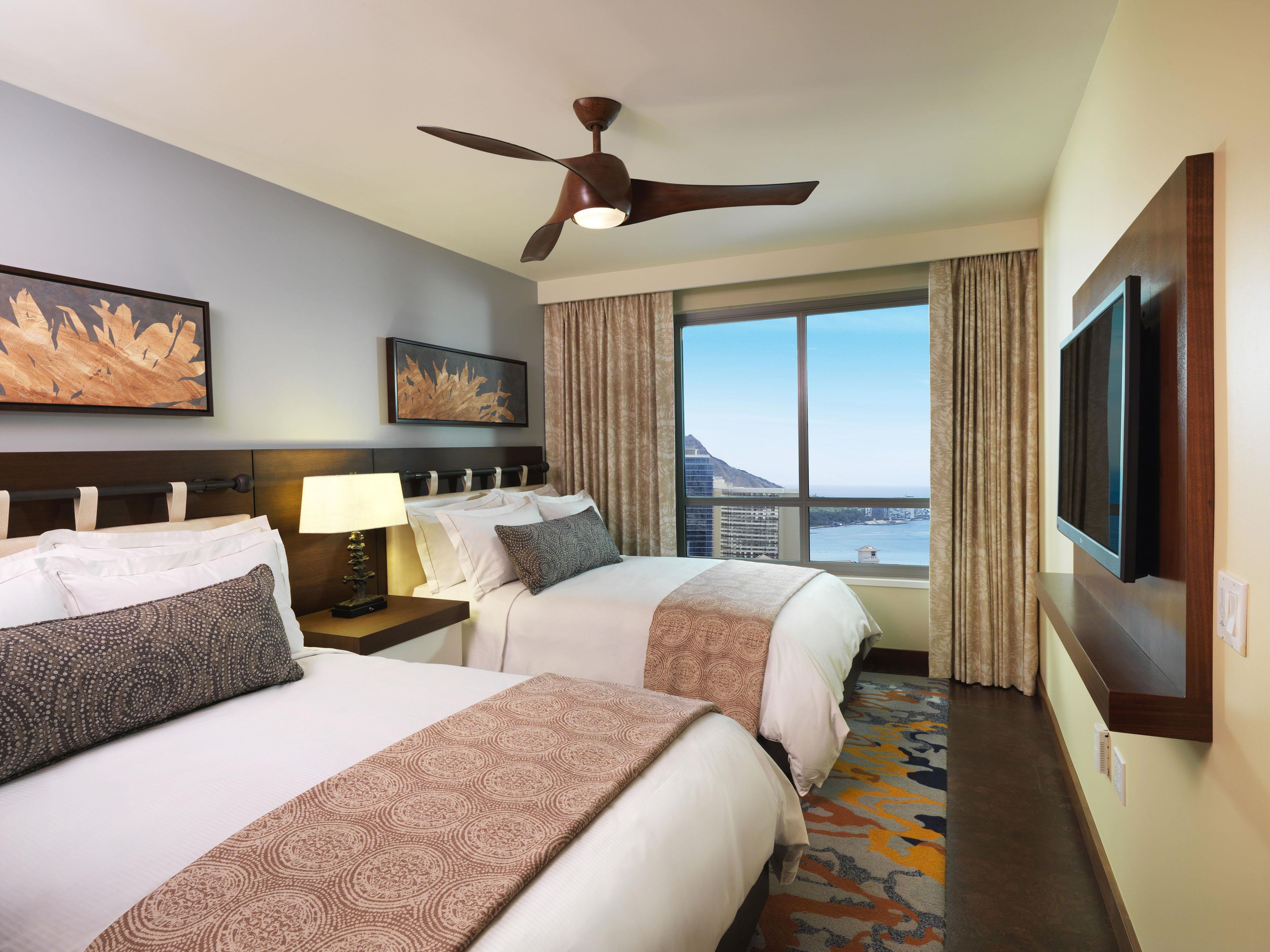 Quarto de hotel com duas camas, ventoinha no teto, janela grande com vista e televisão de tela plana.