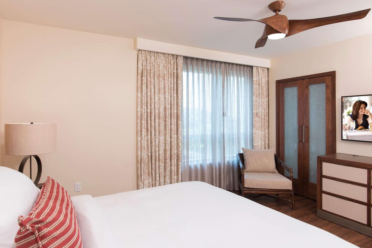 Ein Hotelzimmer mit Bett, Stuhl, Schreibtisch, Deckenventilator und einem großen Fenster mit Vorhängen.