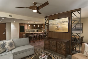 Una sala moderna con un sofá, una mesa de centro y un televisor montado en una pared de madera. Se ve una zona de cocina con taburetes de bar y una mesa de comedor al fondo.