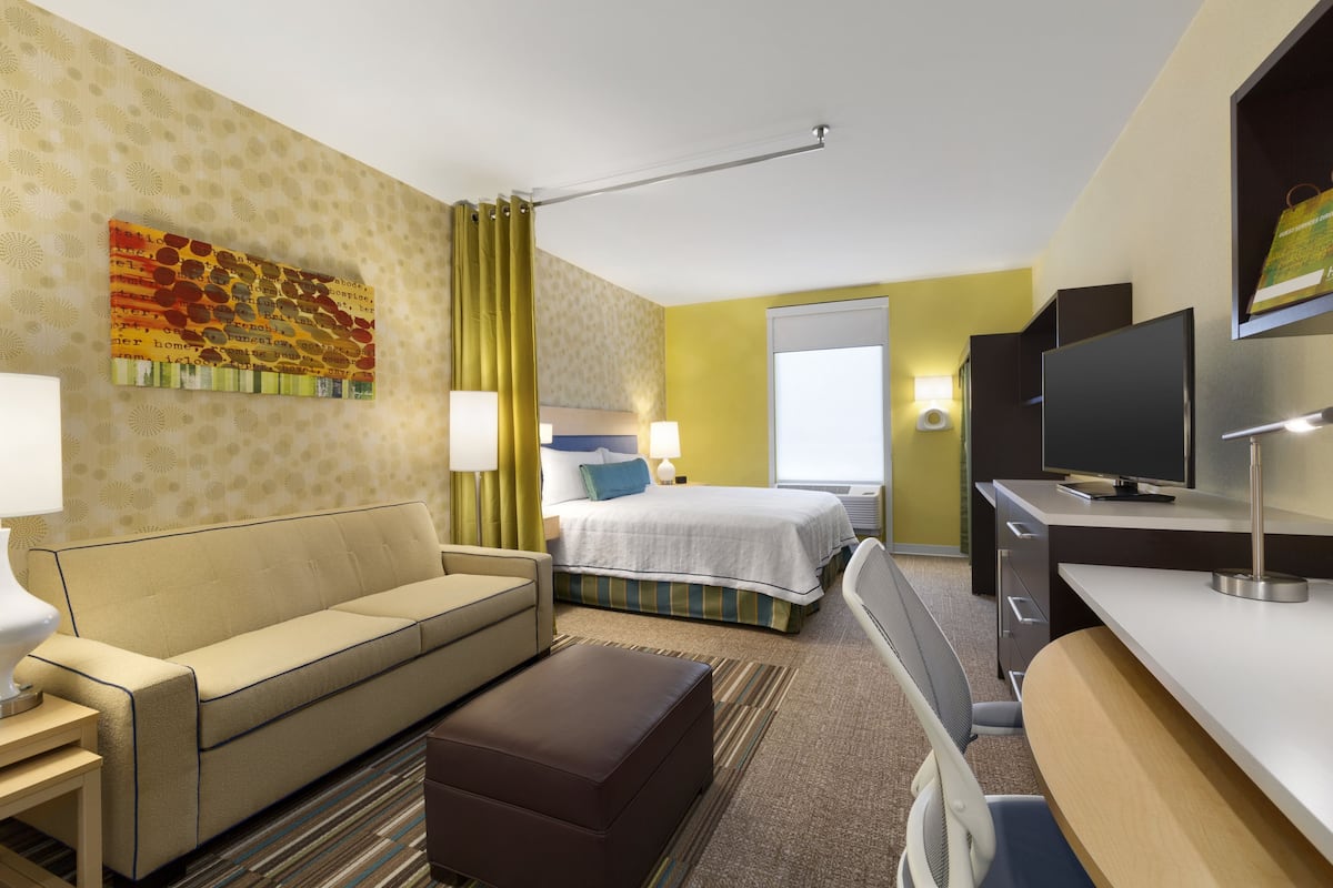 A hotel room with a bed, a sofa, a desk, a television, and a painting on the wall.