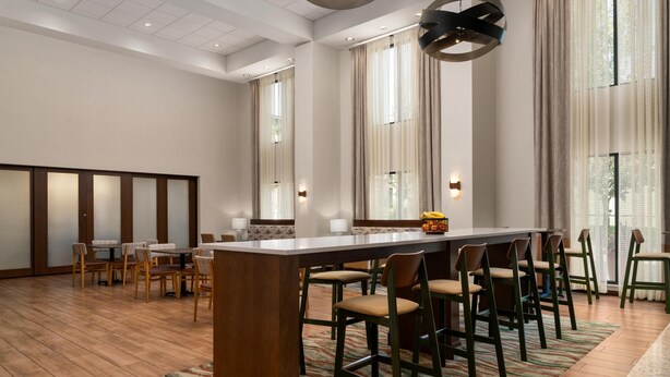 Un hall de hotel espacioso con una zona de bar central, varias opciones de asientos y ventanas grandes que permiten la entrada de luz natural.