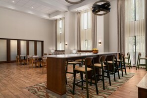 Un hall d’hôtel spacieux doté d’un bar central, de nombreux espaces pour s’asseoir et de grandes fenêtres laissant entrer la lumière naturelle.