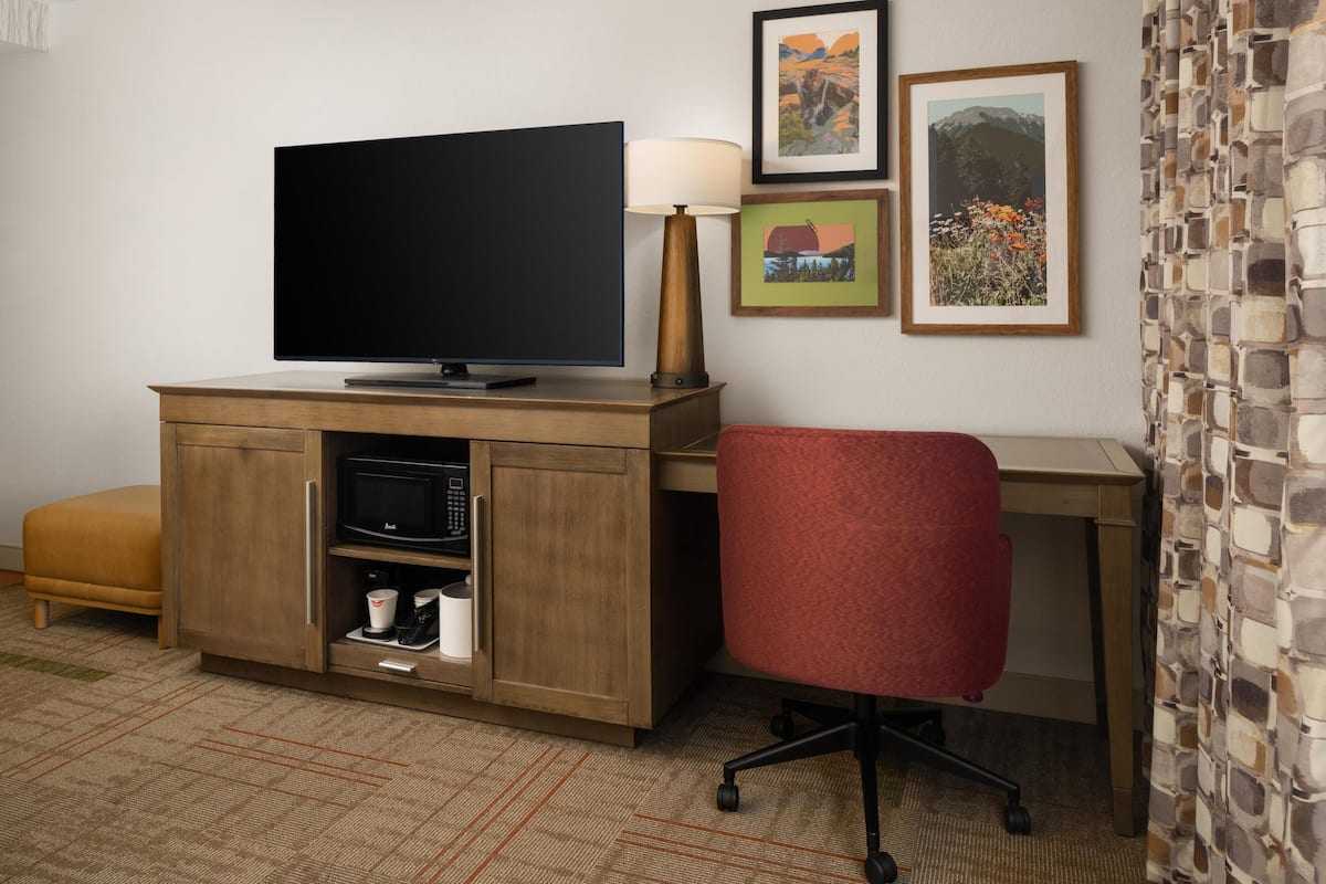 A hotel room with a flat-screen TV on a wooden cabinet, a red office chair, a desk, and a stone-patterned wall.