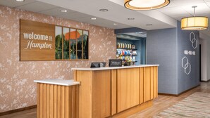 Un mostrador de recepción con un cartel que dice 'Welcome to Hampton', un cartel de 'Hilton Honors' y vistas del área del vestíbulo.