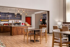 Une chambre d’hôtel moderne avec un plancher en bois, un coin repas avec une table et des chaises, un coin cuisine avec un bar, et une grande fenêtre avec des rideaux.