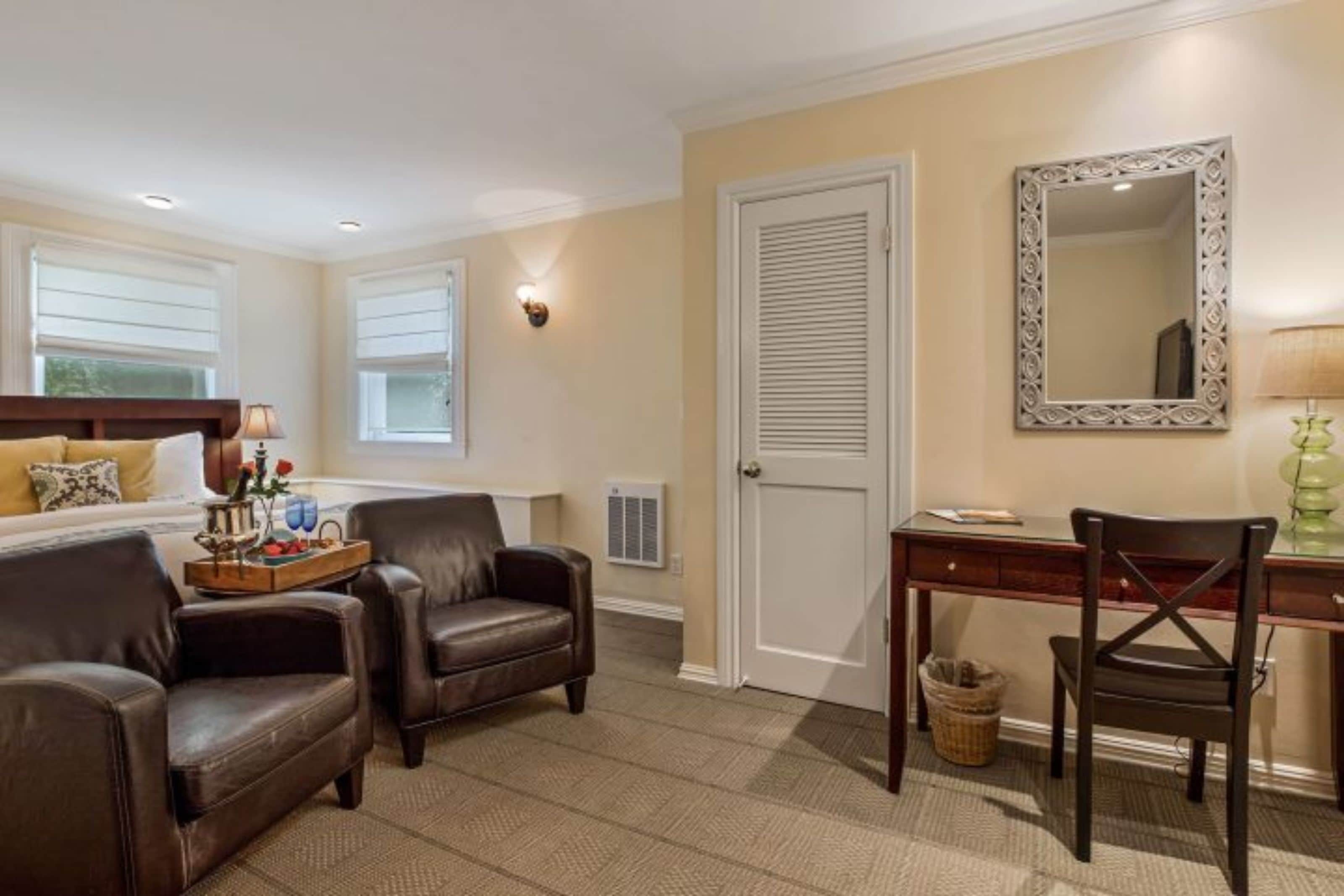 A room with a bed, two leather chairs, a desk, a mirror, and a window with blinds.
