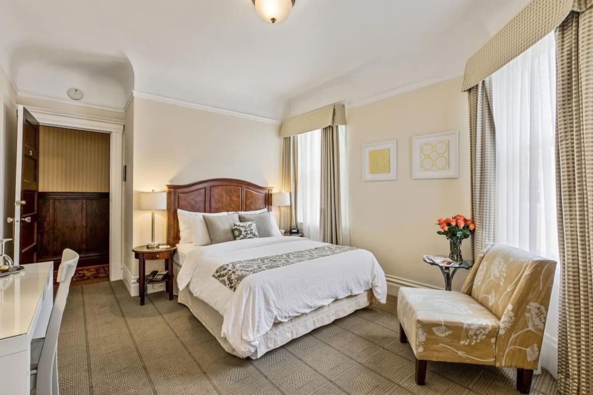 Una camera d'albergo con un letto grande, un comodino con una lampada, una sedia e un tavolino con un vaso di fiori.