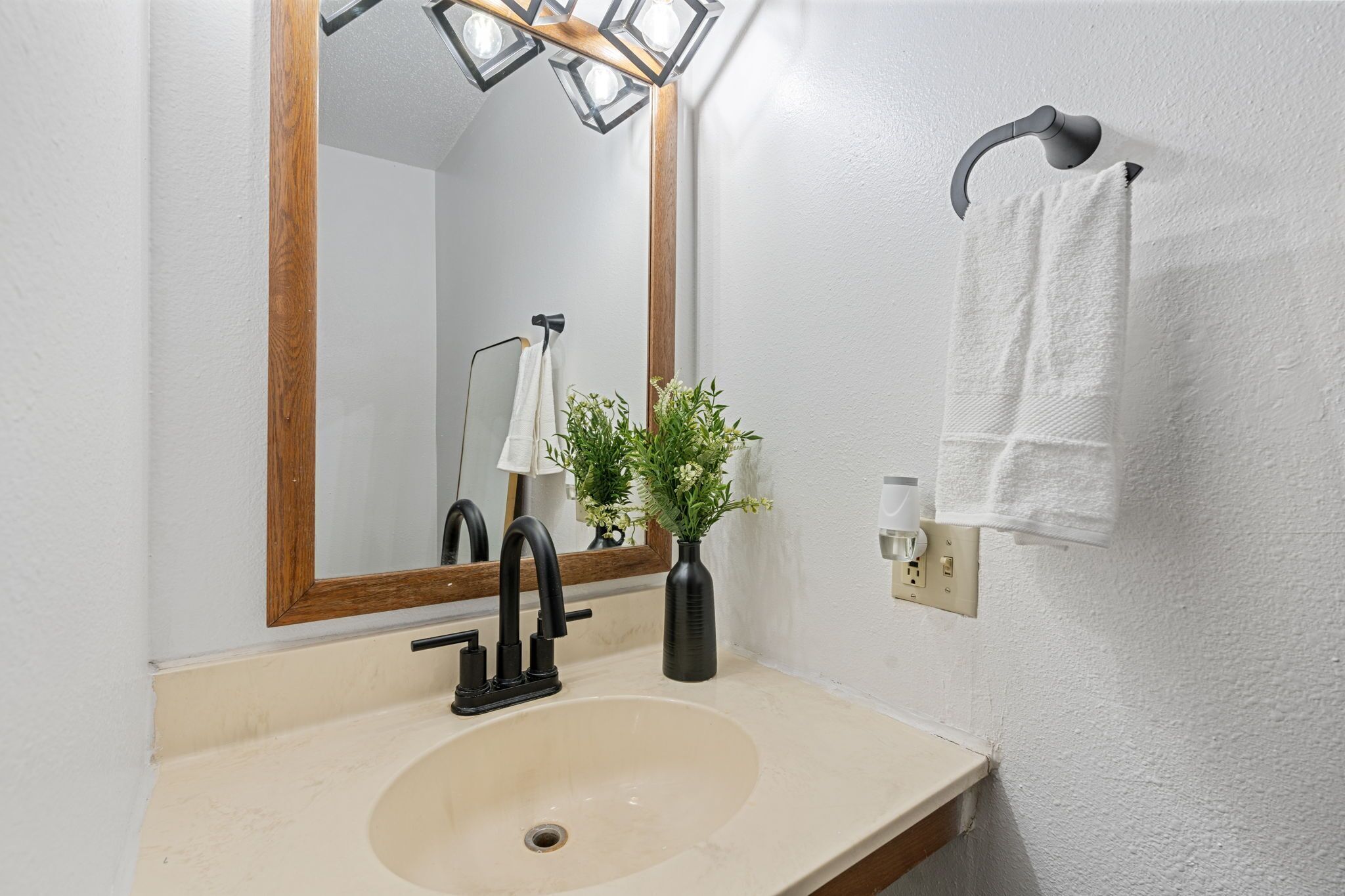 A bathroom with a beige sink, a black faucet, a mirror with a wooden frame, a towel rack with a white towel, and a vase with green plants.