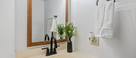 A bathroom with a beige sink, a black faucet, a mirror with a wooden frame, a towel rack with a white towel, and a vase with green plants.