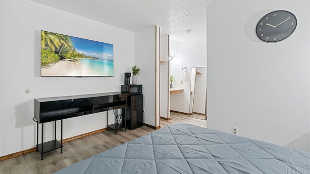 A bedroom with a bed, a desk with a computer, a clock, and a painting of a beach.