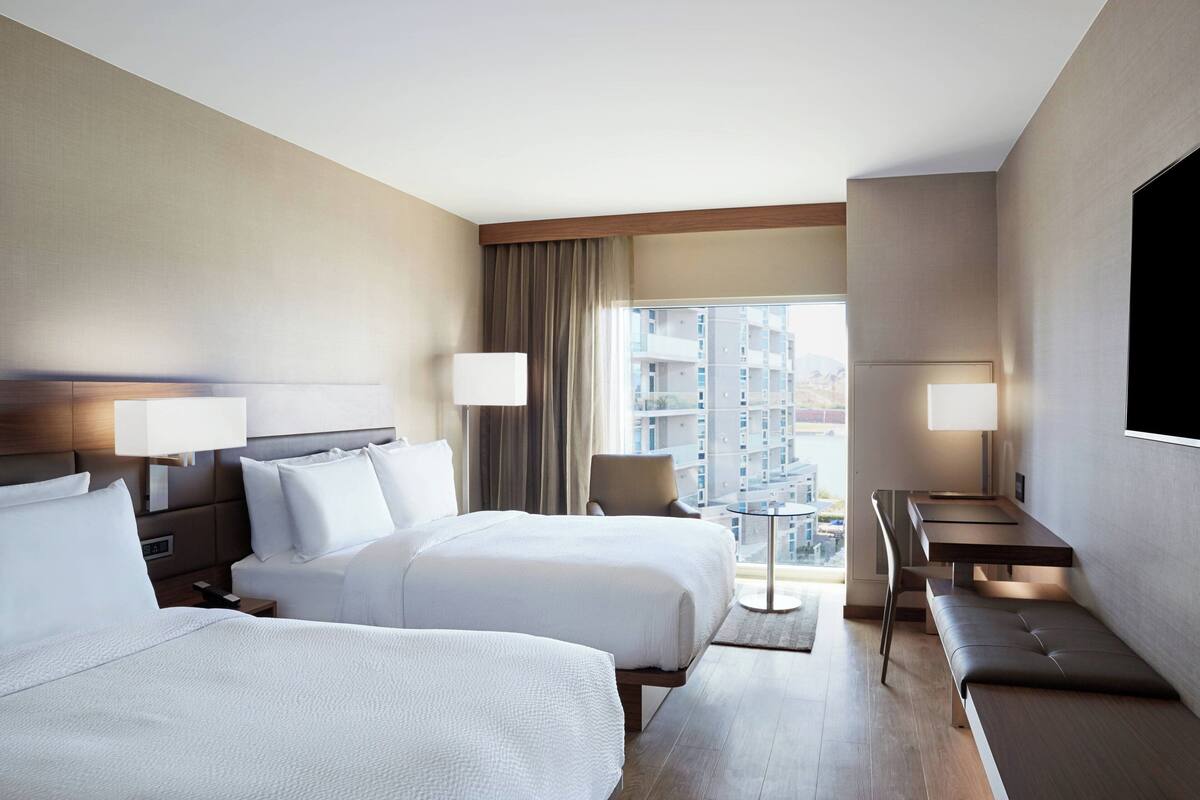 A hotel room with two beds, a desk, and a view of the city.