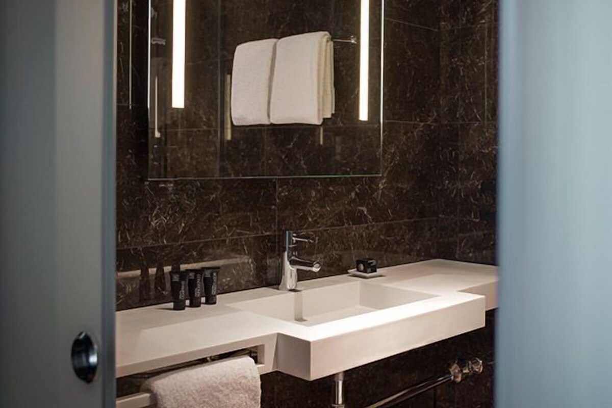 Un baño moderno con un lavamanos blanco, paredes de mármol oscuro y un espejo grande.