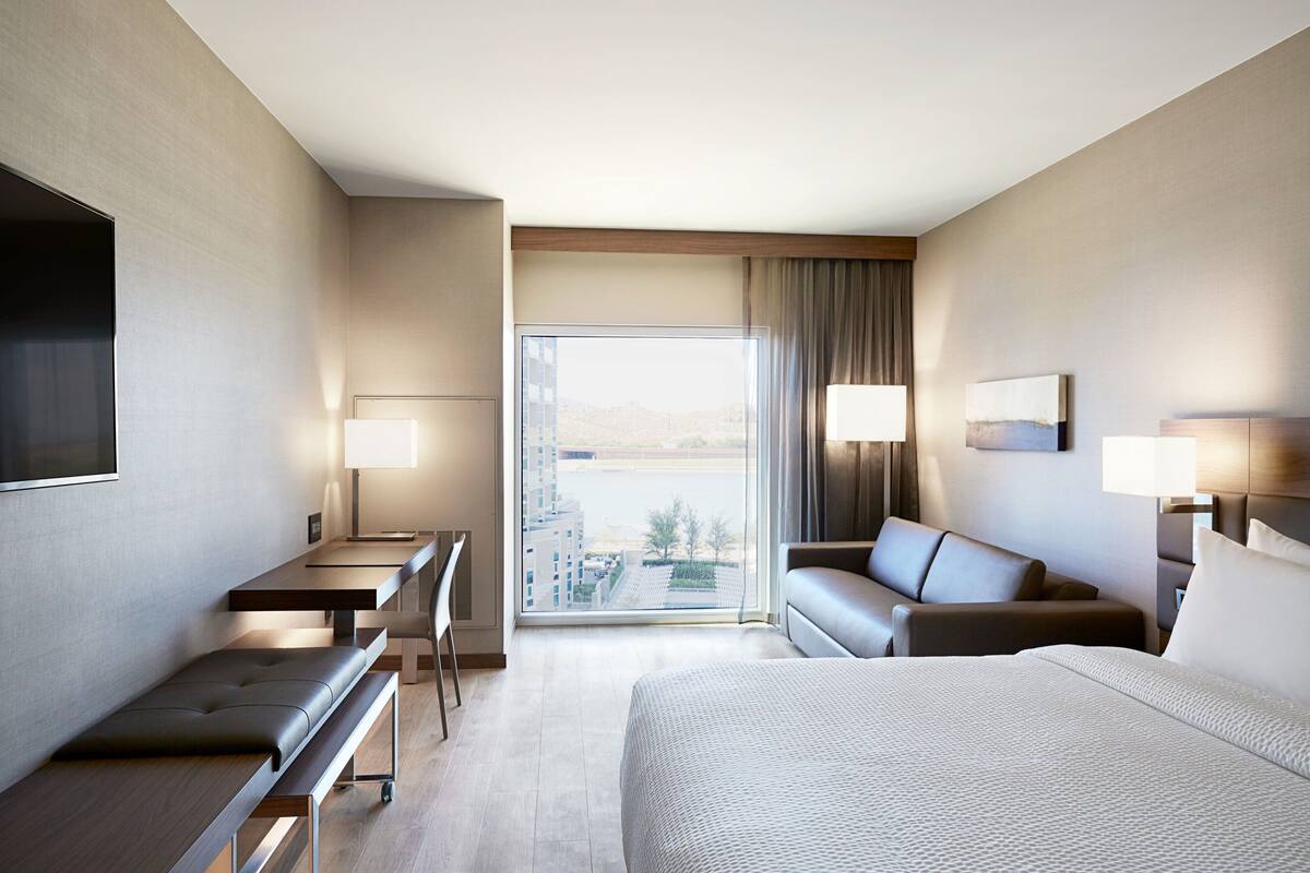 A modern hotel room with a large bed, a desk with a chair, a sofa, and a view of the city through a large window.
