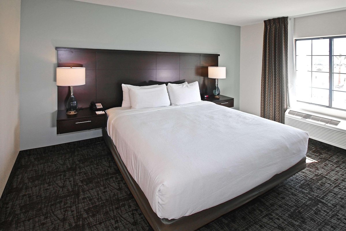 A hotel room with a large bed, two bedside tables with lamps, a window with curtains, and a wall-mounted air conditioning unit.