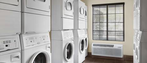 Un cuarto de lavandería con lavadoras y secadoras apiladas, una ventana con persianas y una rejilla de ventilación.