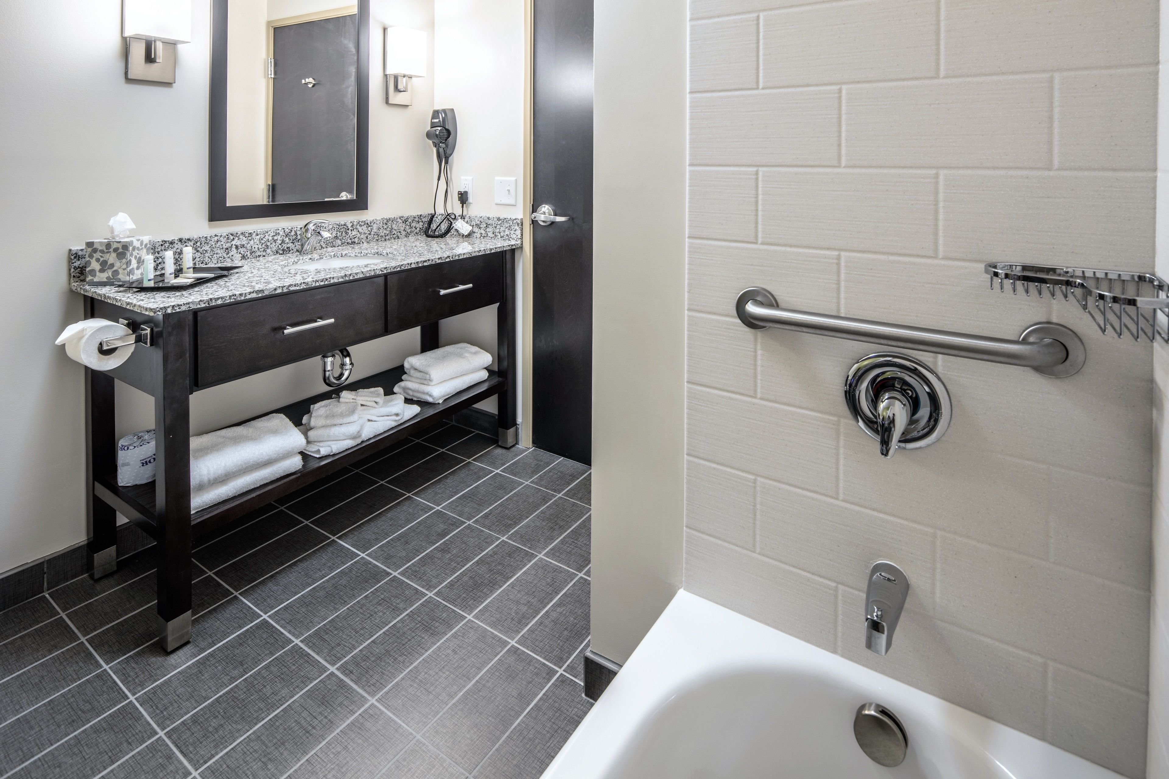 A modern bathroom with a granite countertop sink, a mirror, a towel rack, and a shower with a grab bar.