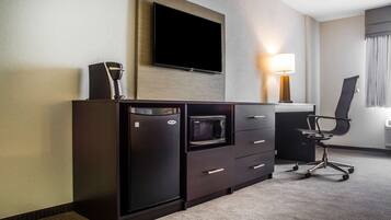 A hotel room with a flat-screen TV mounted on the wall, a microwave, a refrigerator, a desk with a lamp, and a chair.