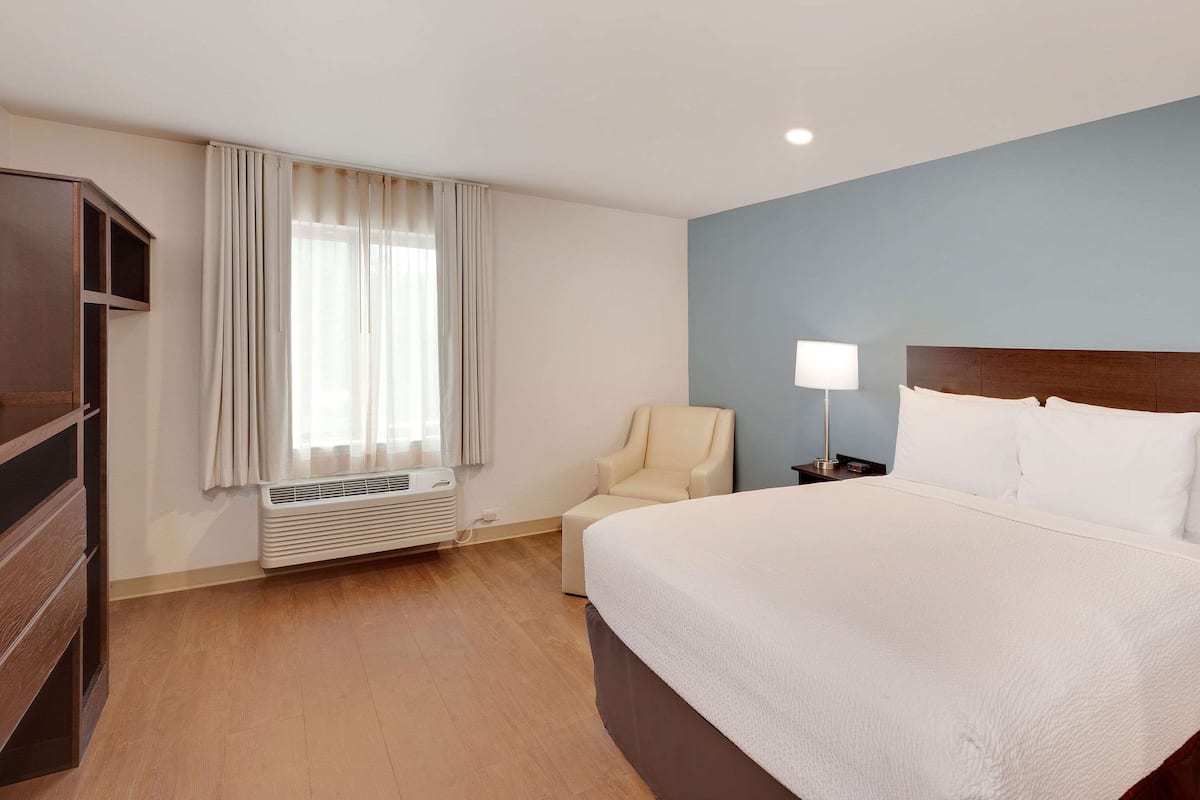 A hotel room with a large bed, a chair, a desk with a lamp, and a wardrobe.