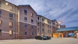A multi-story hotel with a brick facade, a covered entrance, and a parking lot with cars. - WoodSpring Suites Las Colinas - Northwest Dallas (Irving)
