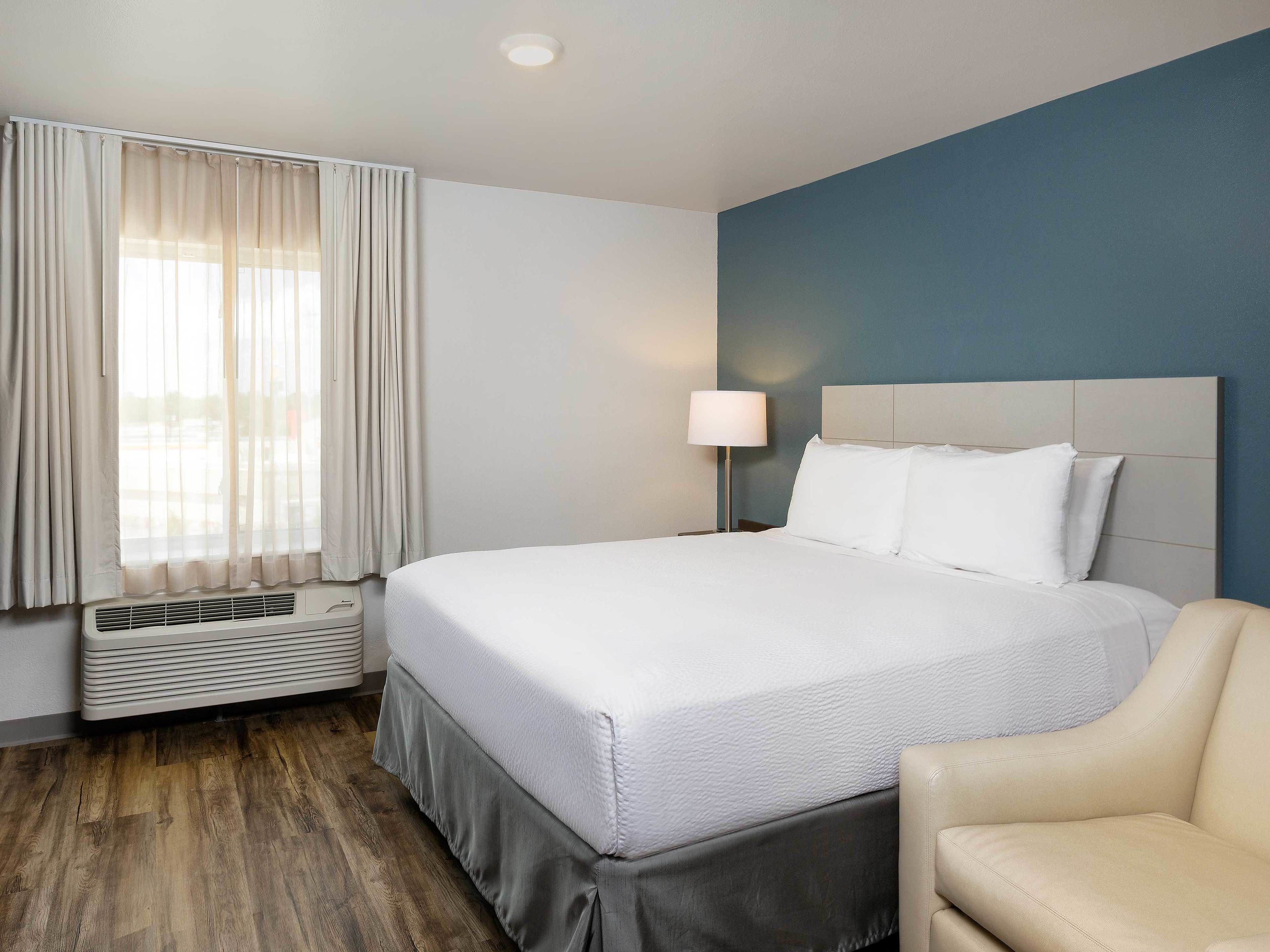 Habitación de hotel con cama, una silla, una mesita de noche con lámpara y ventana con cortinas.
