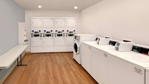 A laundry facility with multiple washing machines and dryers, a bench, and a wall-mounted control panel. - WoodSpring Suites Las Colinas - Northwest Dallas (Irving)