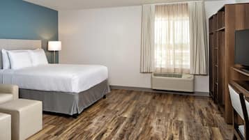 Una habitación de hotel con cama, escritorio, silla, televisor, ventana con cortinas y aire acondicionado instalado en la pared.