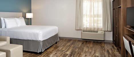 A hotel room with a bed, a desk, a chair, a TV, a window with curtains, and a wall-mounted air conditioner.
