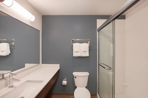 A modern bathroom with a large mirror, two sinks, a toilet, and a glass shower enclosure. - WoodSpring Suites Houston IAH Airport (Humble)