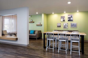 A dining area with a wooden table and four chairs, a sofa, and a window with sheer curtains. - WoodSpring Suites Houston IAH Airport (Humble)