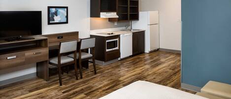 A compact hotel room with a wooden floor, a desk with a chair, a kitchenette with a refrigerator, and a television.