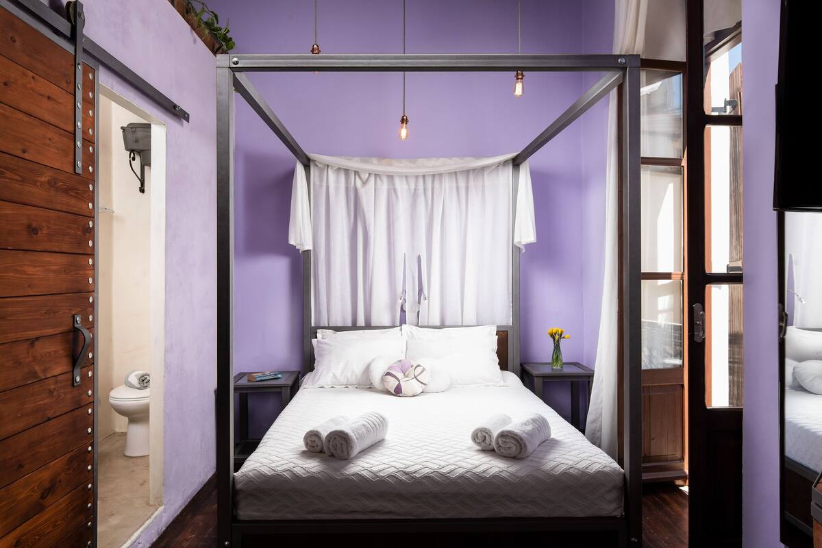 A four-poster bed with white linens and pillows, a bedside table with a vase of flowers, and a view of the outside through a window.