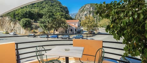 Un balcón con mesa y sillas, con vista a una calle con coches aparcados y un edificio de techo rojo.