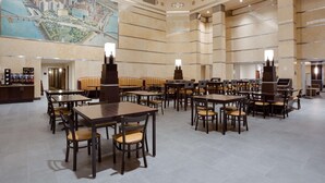 A spacious hotel lobby with multiple dining tables and booths, a large cityscape mural on the wall, and a high ceiling with decorative moldings. - Drury Plaza Hotel Pittsburgh Downtown (Pittsburgh)