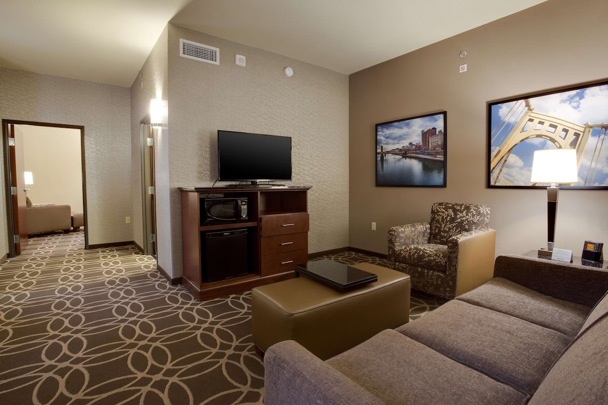 A hotel room with a flat-screen TV, a sofa, a chair, a small table, and a carpeted floor.